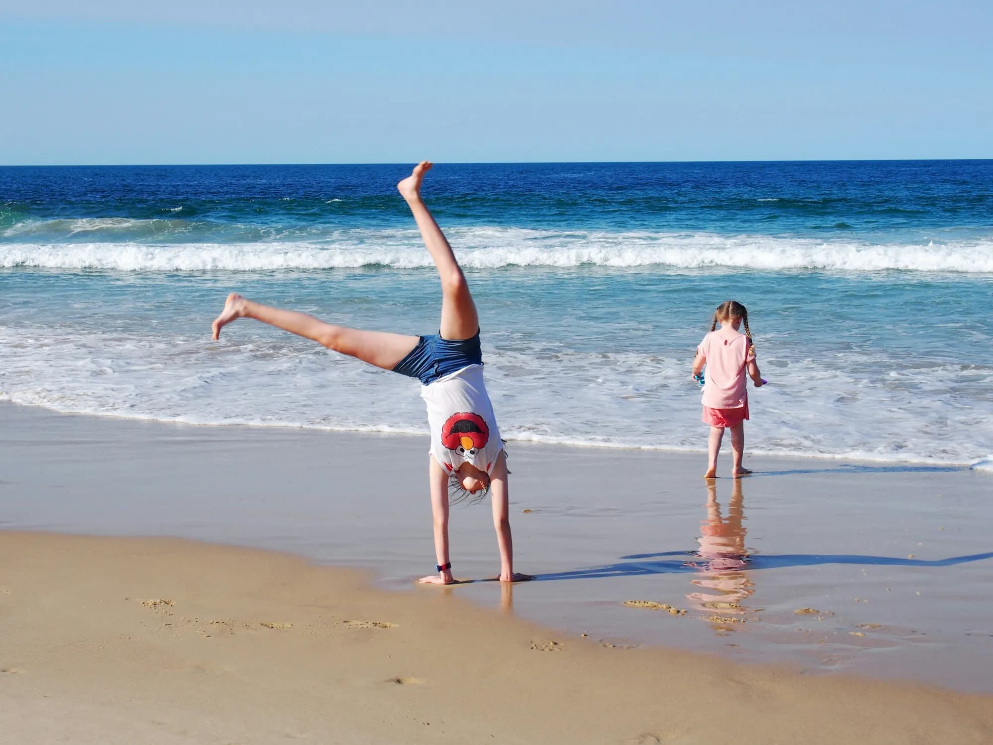 Two children at the edge of the surf on a Gold Coast beach, one mid-cartwheel on the wet sand