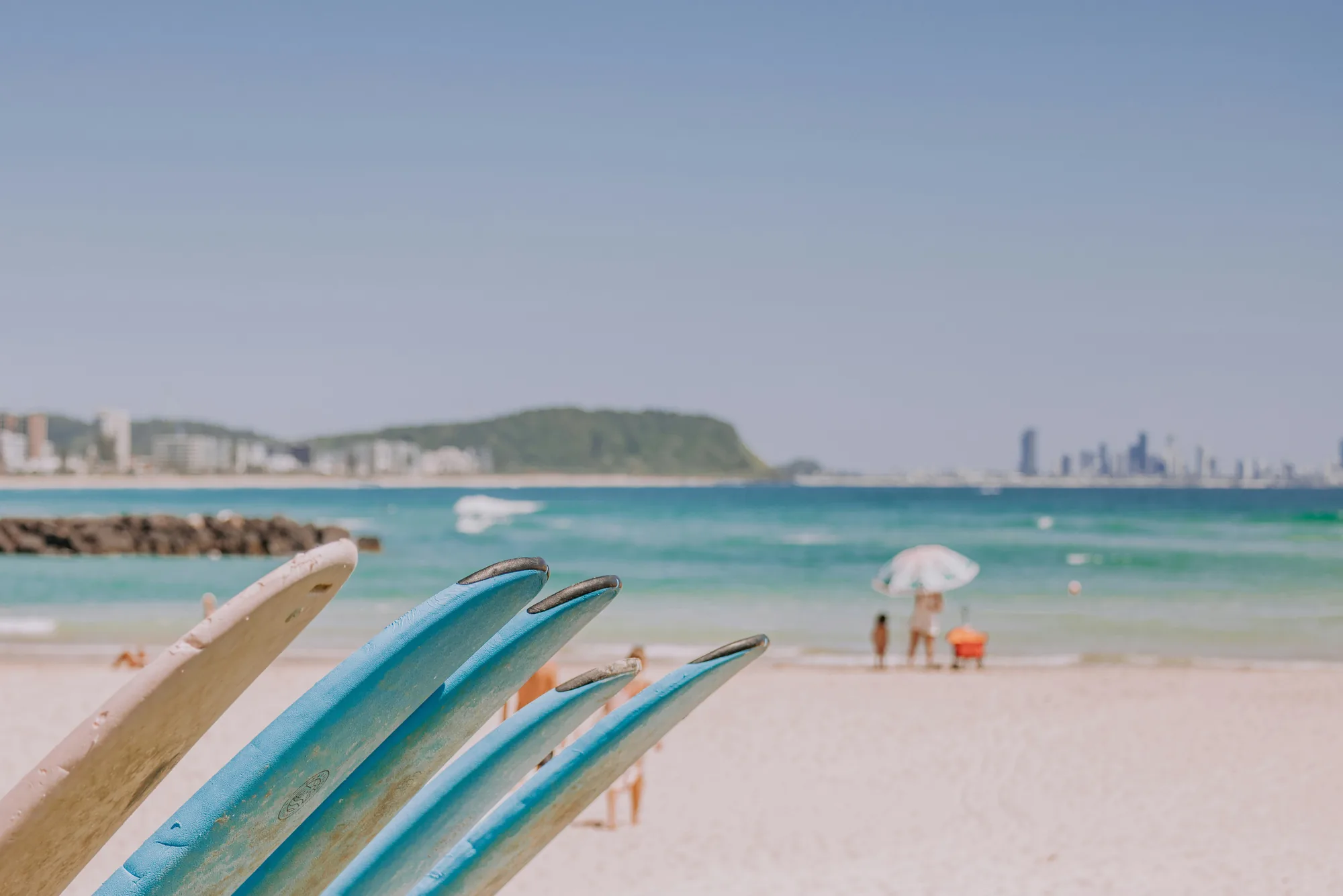 Foam surfboards standing on the sand at Burleigh, with a family at the water, the headland behind and the coast stretching north