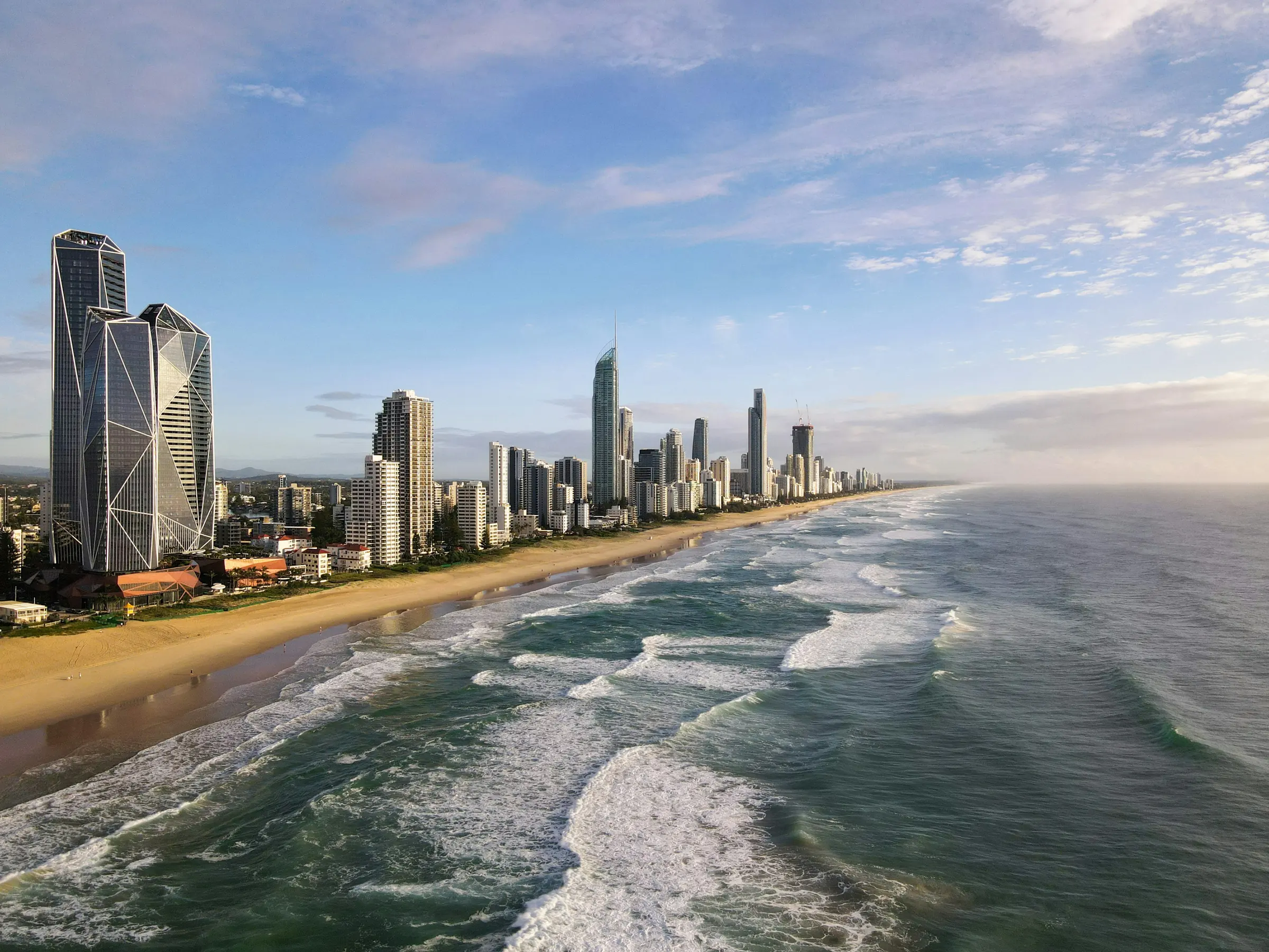 The Gold Coast shoreline from the air in morning light, with surf rolling onto the beach and the towers of Surfers Paradise along the sand