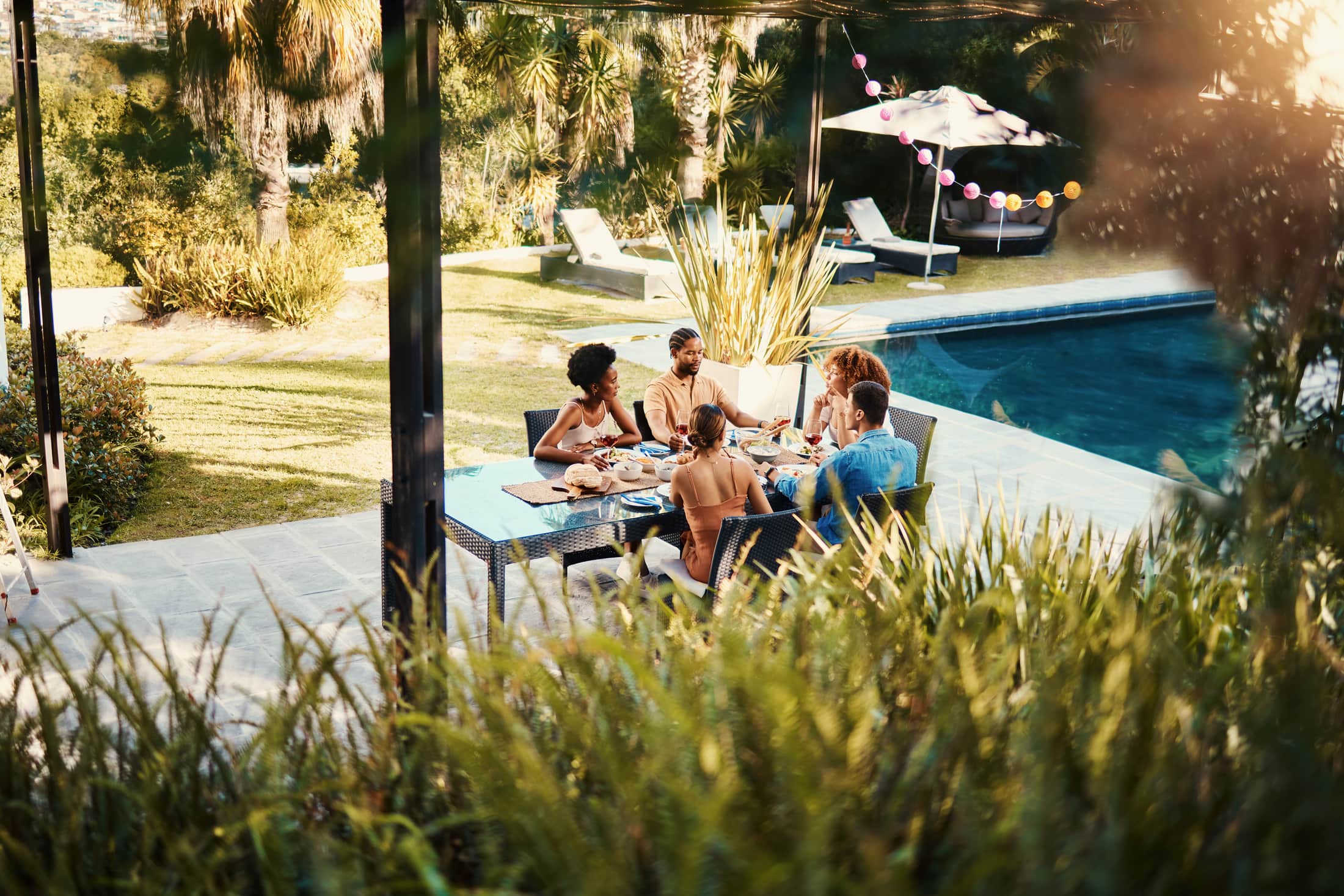 Friends sharing lunch at an outdoor table beside a pool in a garden, seen through tall grasses