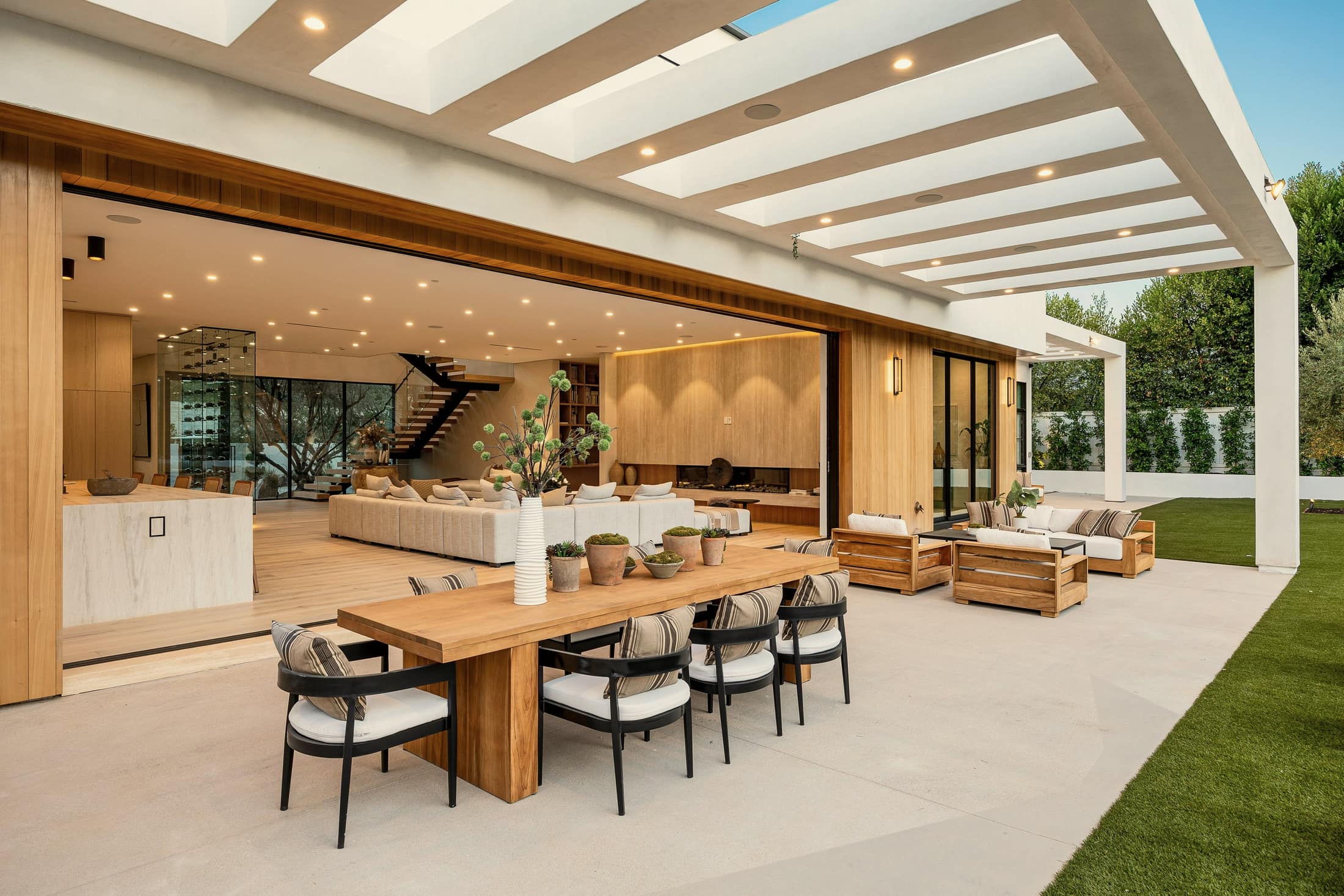 A living room opening onto a covered terrace, with a timber dining table, outdoor seating and a lawn beyond