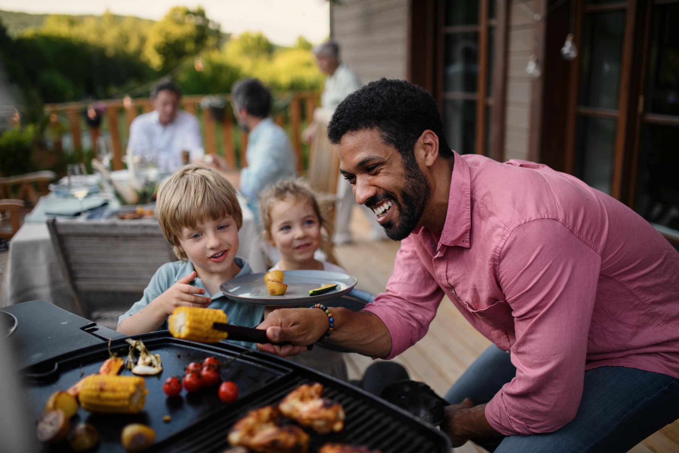 A father at the barbecue on a timber deck, serving two children while the rest of the family sits at the table behind